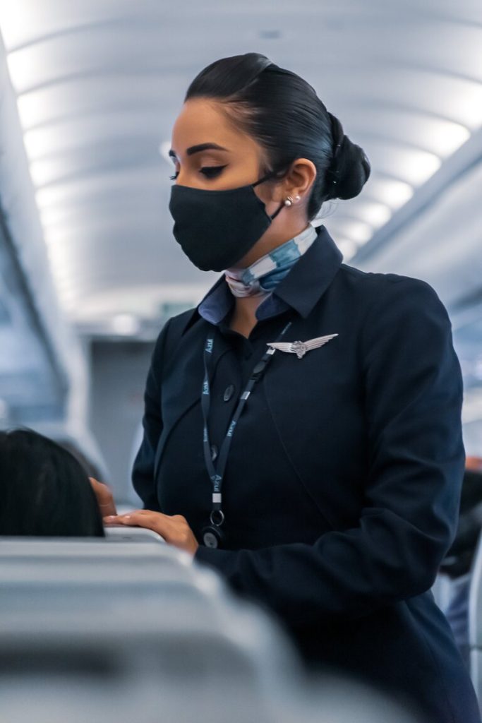 masked-female-flight-attendant-resting-hand-on-passenger-head-rest